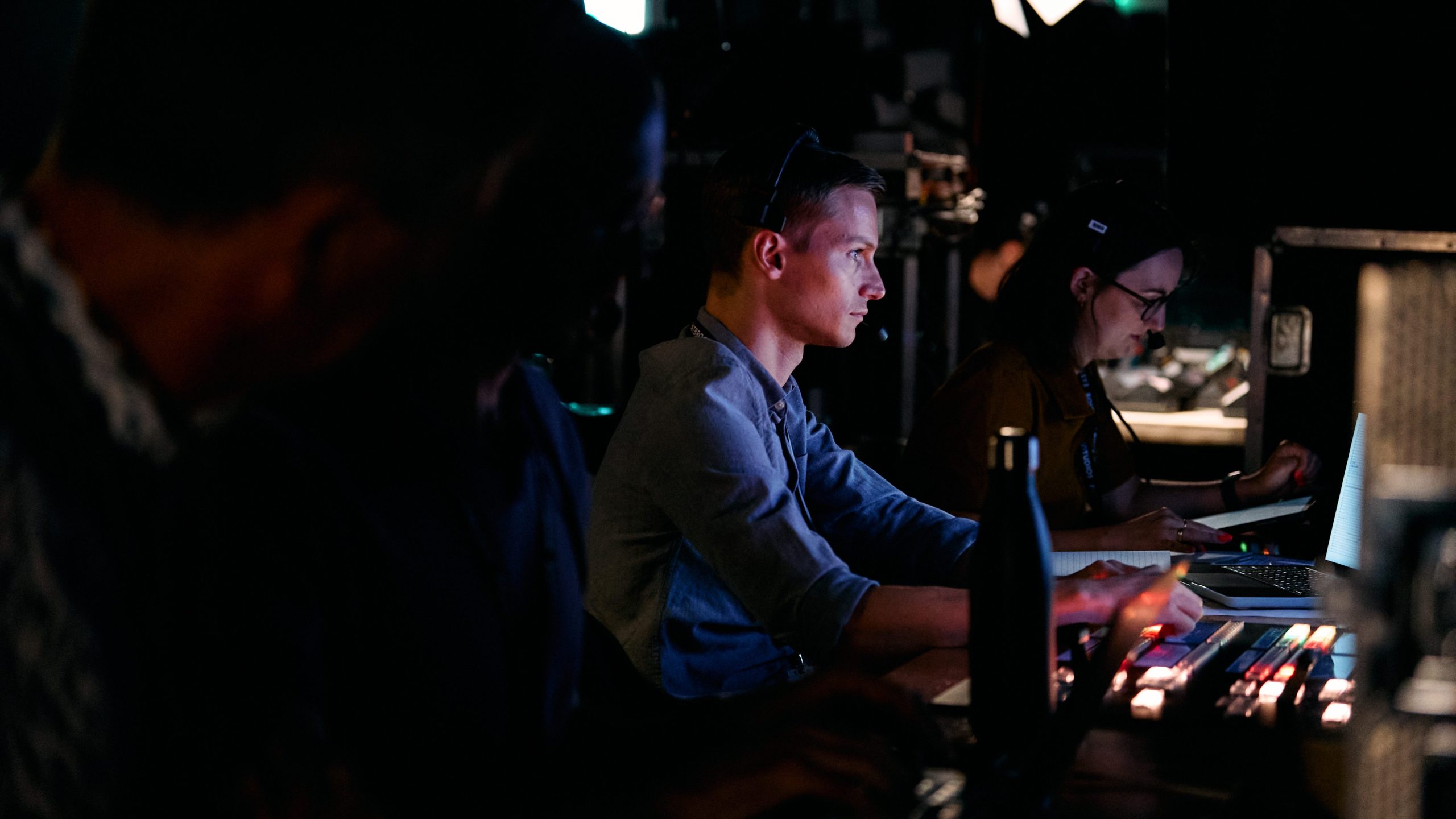 A profile view of people seated in a dimly lit television broadcast gallery, with faces lit by screens.